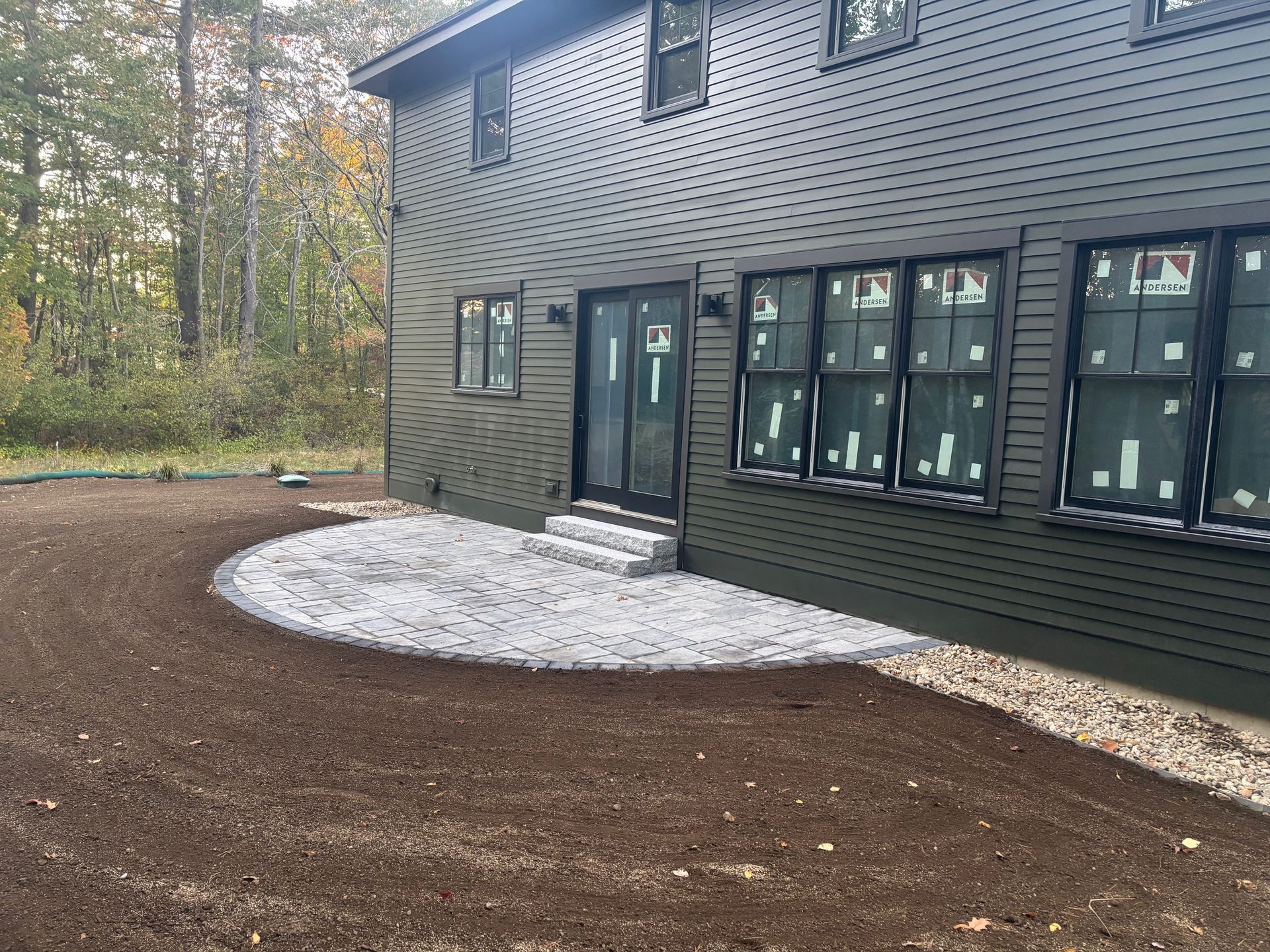 Gray gravel patio area next to a dark-sided house with new windows and a wooded backdrop.