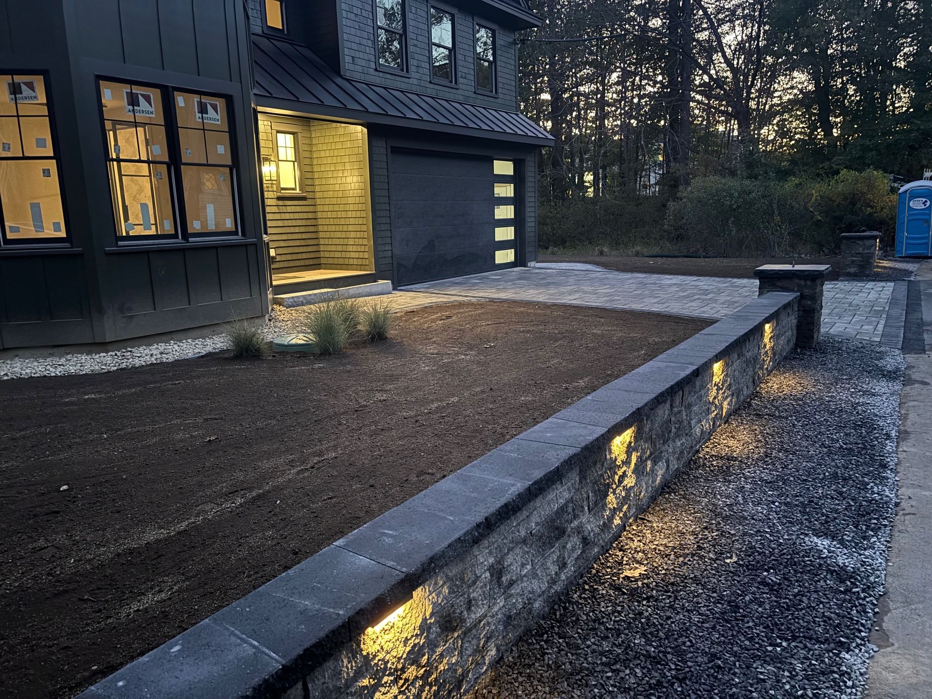 Modern dark house exterior with lit stone retaining wall and gravel driveway.