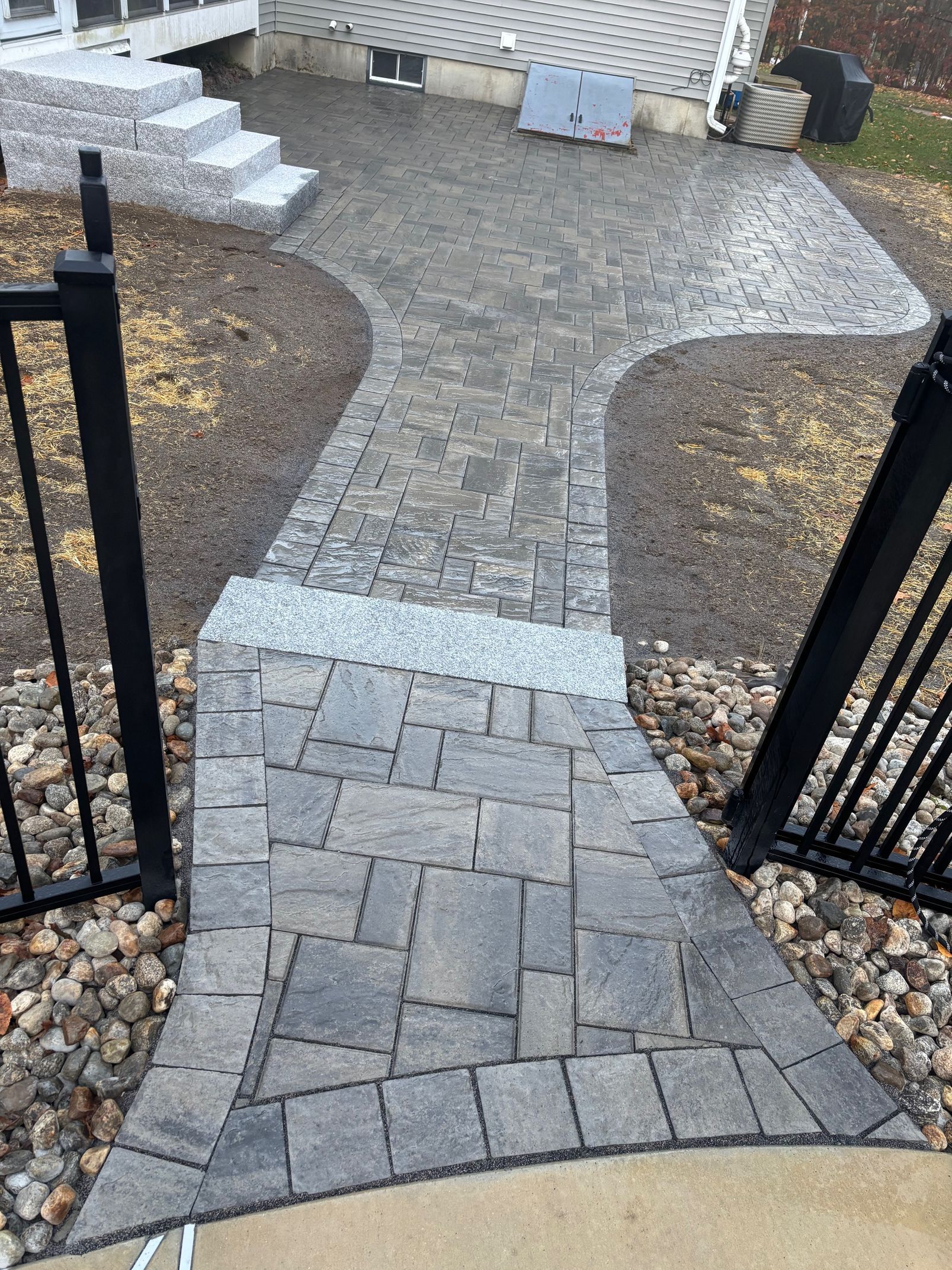 Stone walkway with steps leading to a yard, featuring dark grey pavers and a black fence.