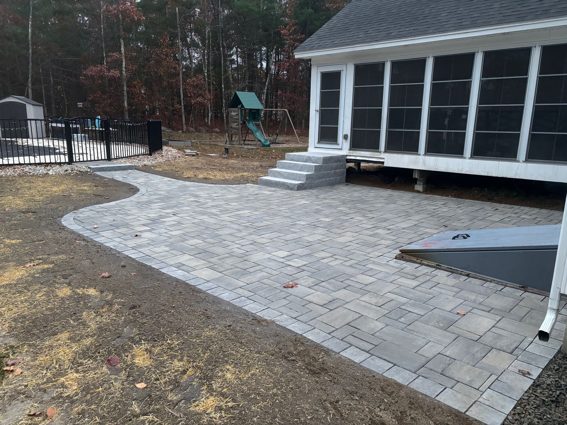 Brick patio next to a house with screened porch and stairs, with a pathway to a fence and playground in the yard.