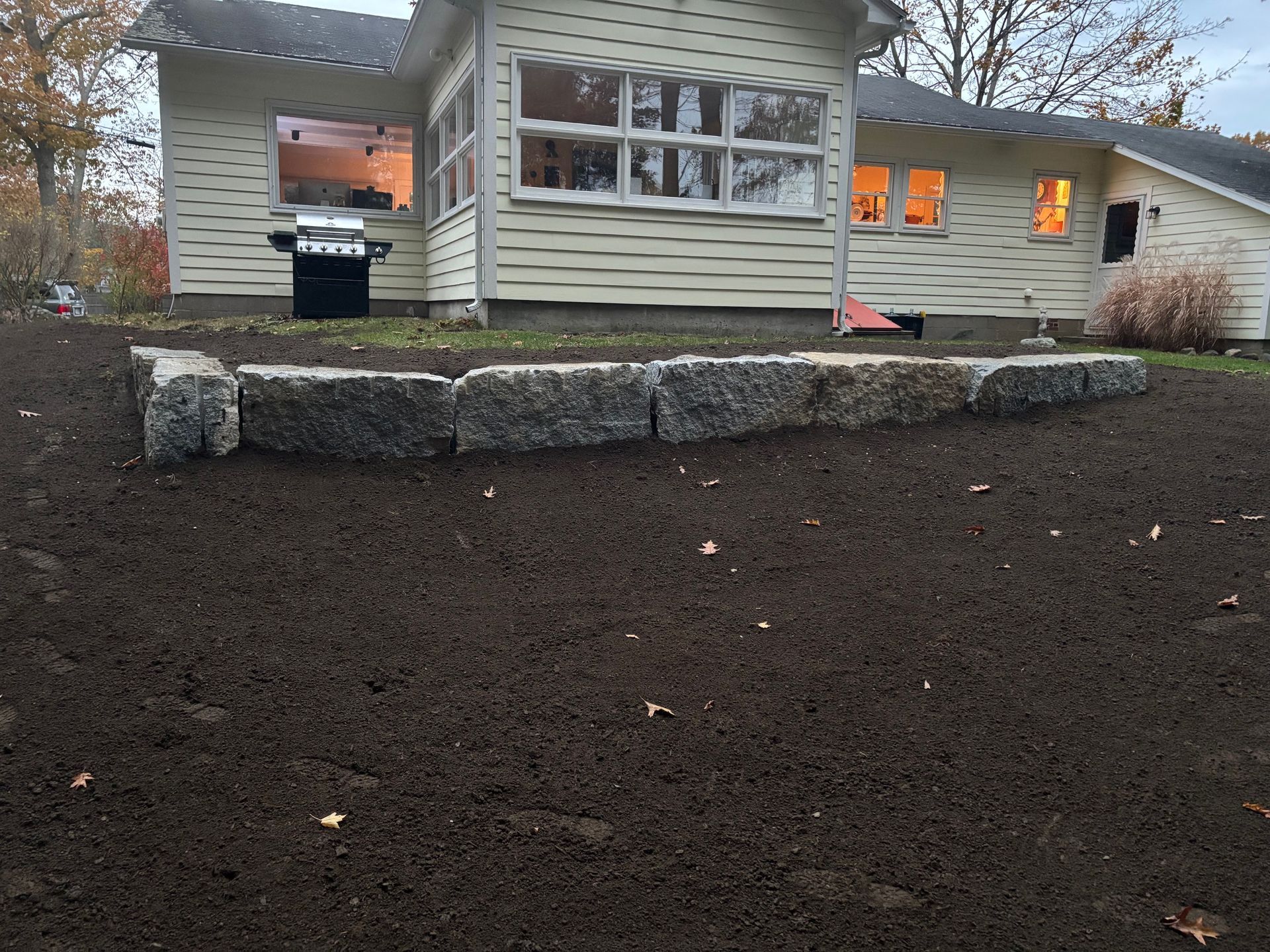 Granite retaining wall in front of a light-colored house with windows; freshly tilled soil.