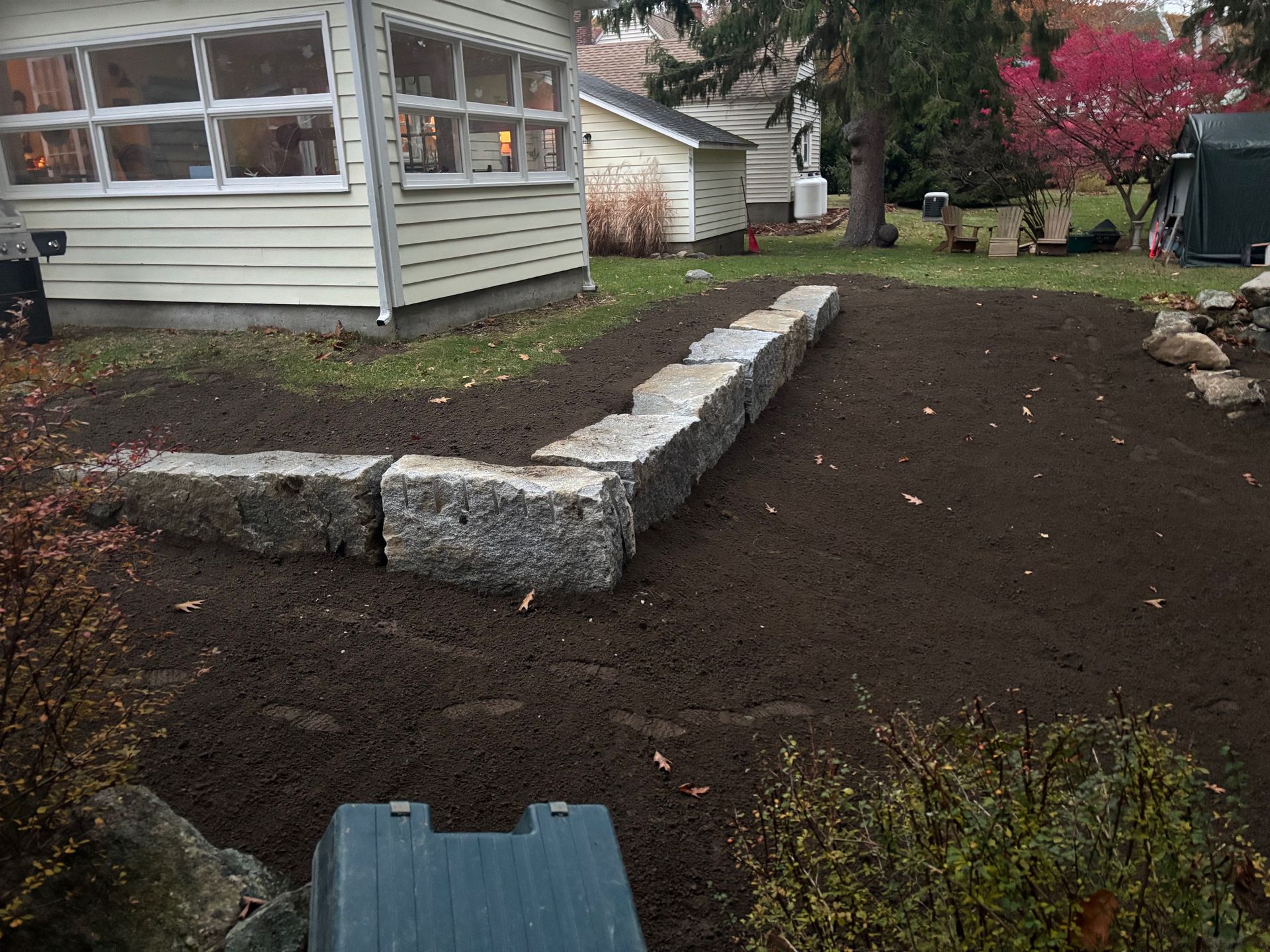 Stone retaining wall in a yard, newly covered with dark soil, beside a light-colored house.