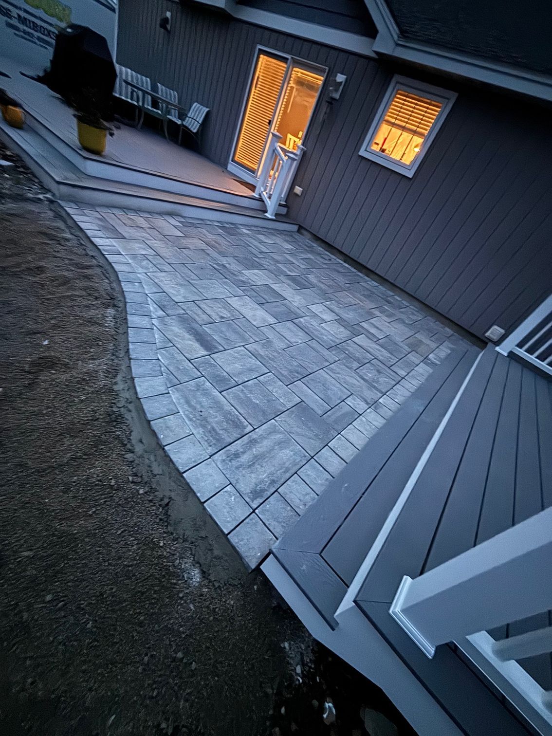 Stone patio next to a gray house, connecting to a composite deck. Evening, warm interior lights.