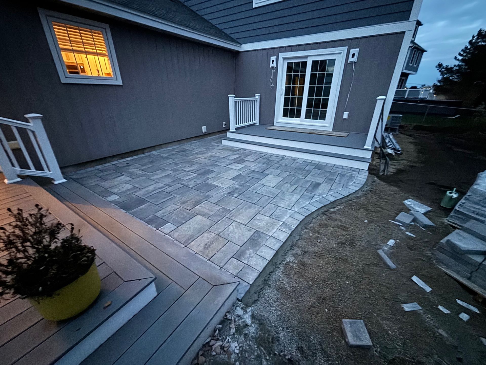 Gray brick patio next to a gray composite deck, leading to a sliding door. Exterior of a house.