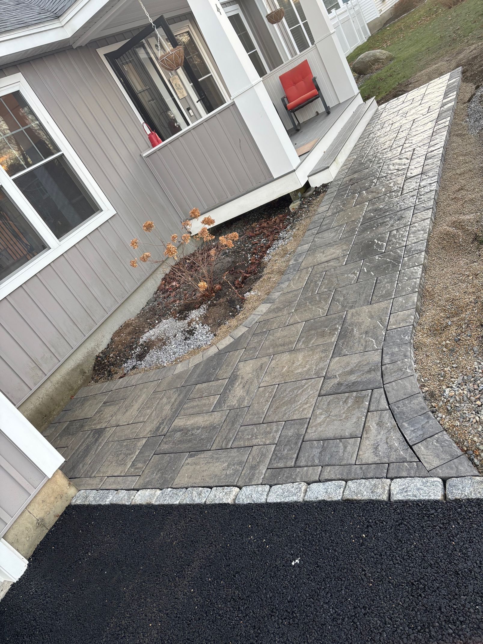 A paved pathway with a dark border leads to a house with a porch and red chair.