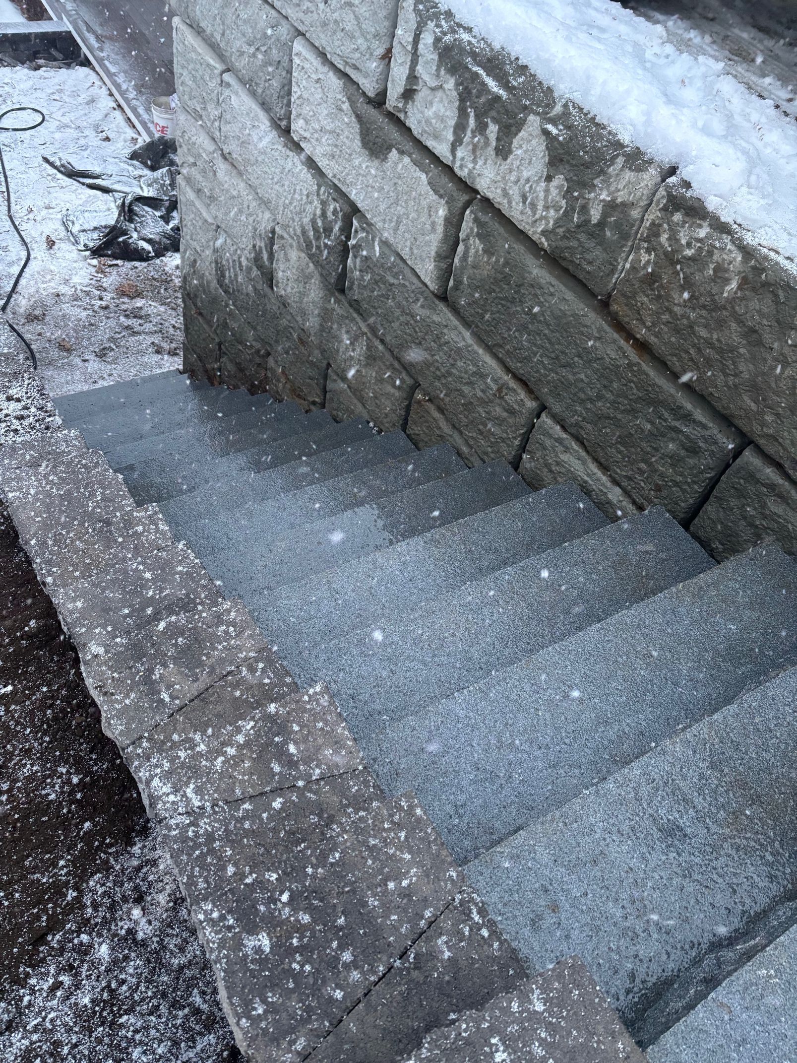 Stone steps leading down, next to a stone wall. Snow flurries on the steps and wall.