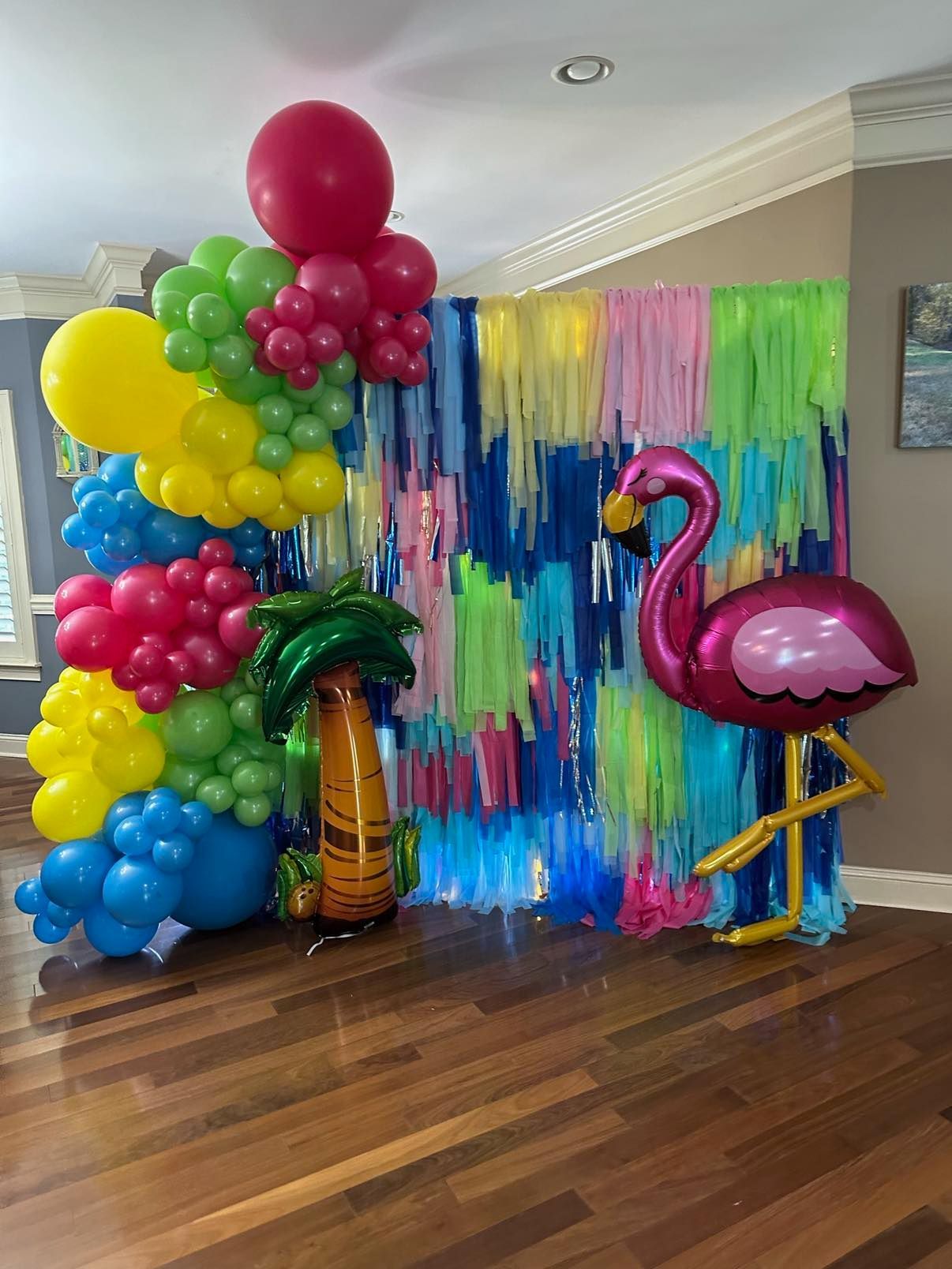 Tropical party scene: flamingo balloon, palm tree balloon, colorful balloon arch, and fringed backdrop on a wooden floor.