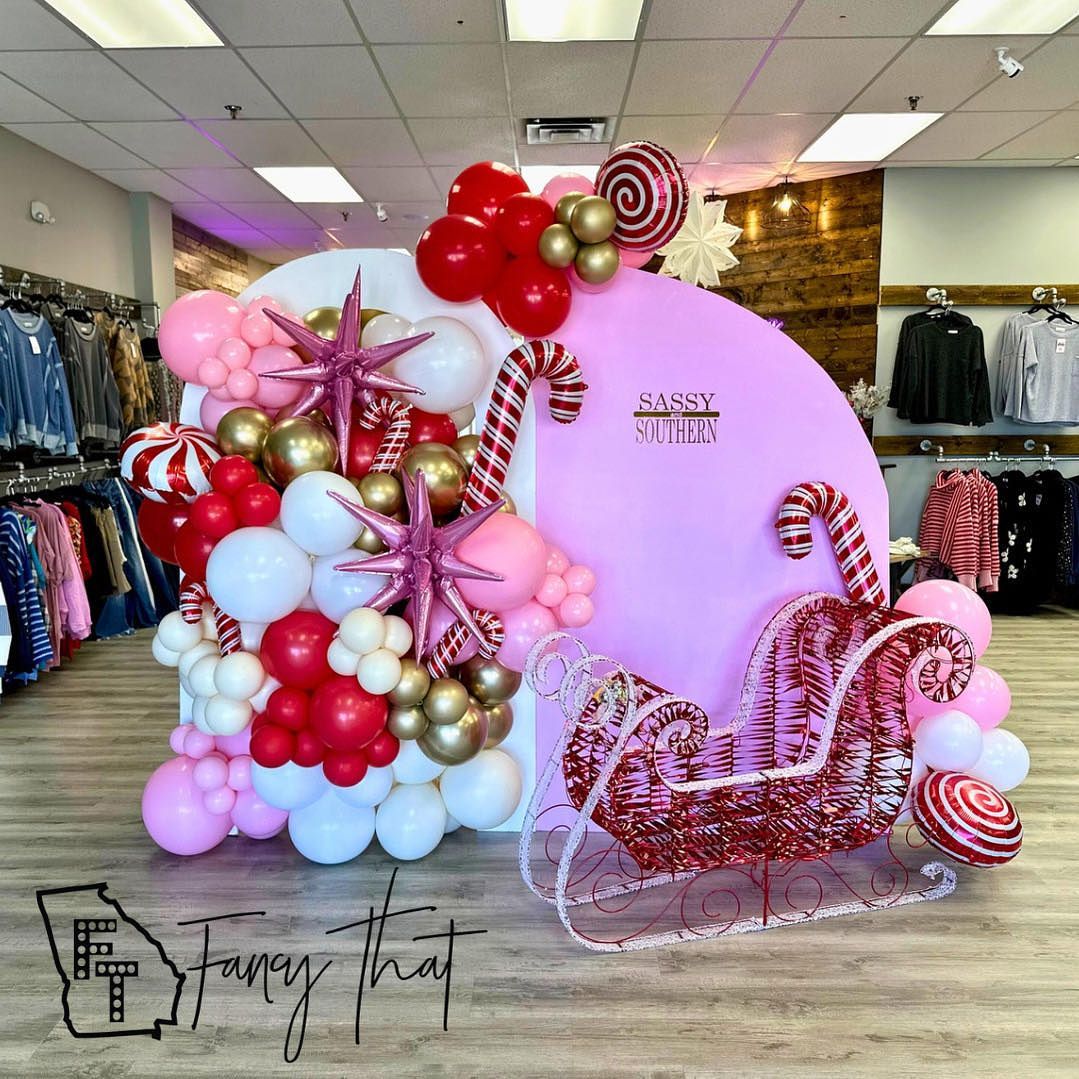 Holiday balloon arch: pink, white, gold, and red balloons with a sleigh, candy canes, and stars.