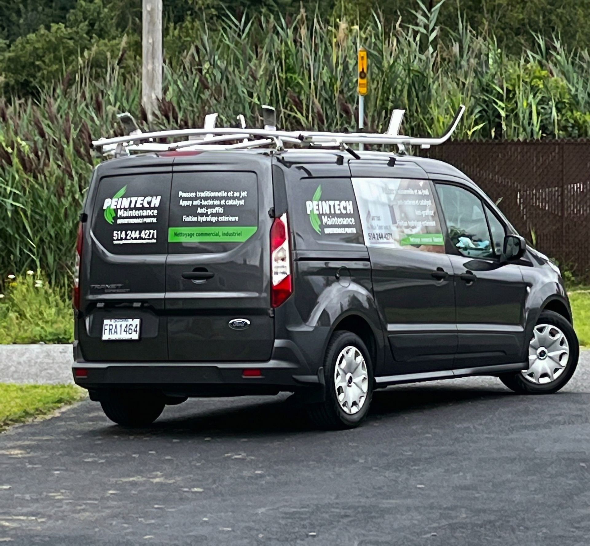 Fourgonnette de service noire stationnée dans une rue, avec des panneaux publicitaires verts et blancs de l'entreprise sur les vitres arrière.