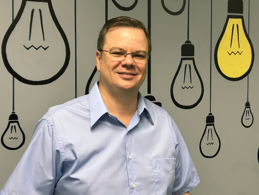 A man wearing glasses is standing in front of a wall with light bulbs painted on it.