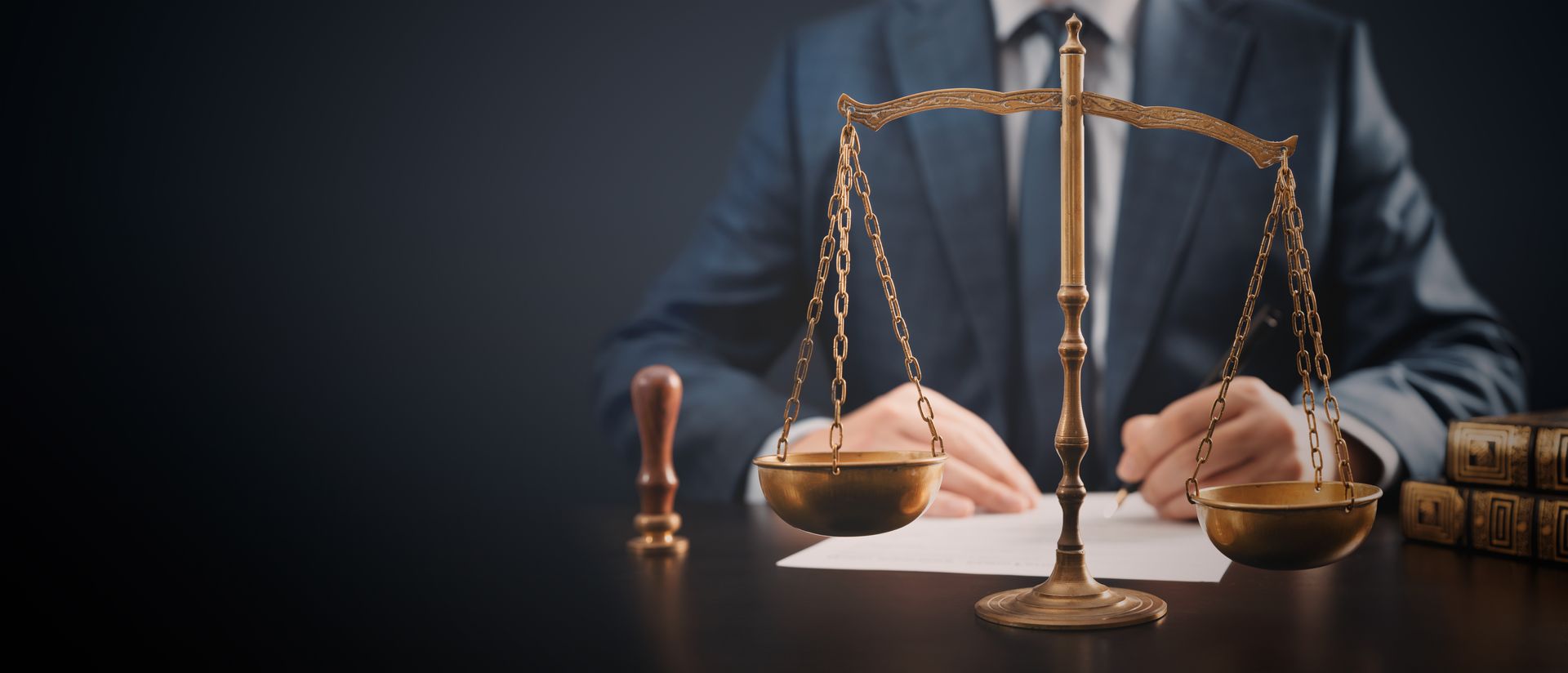 A person in a suit sits behind a desk. A golden scale of justice and books are in the foreground.