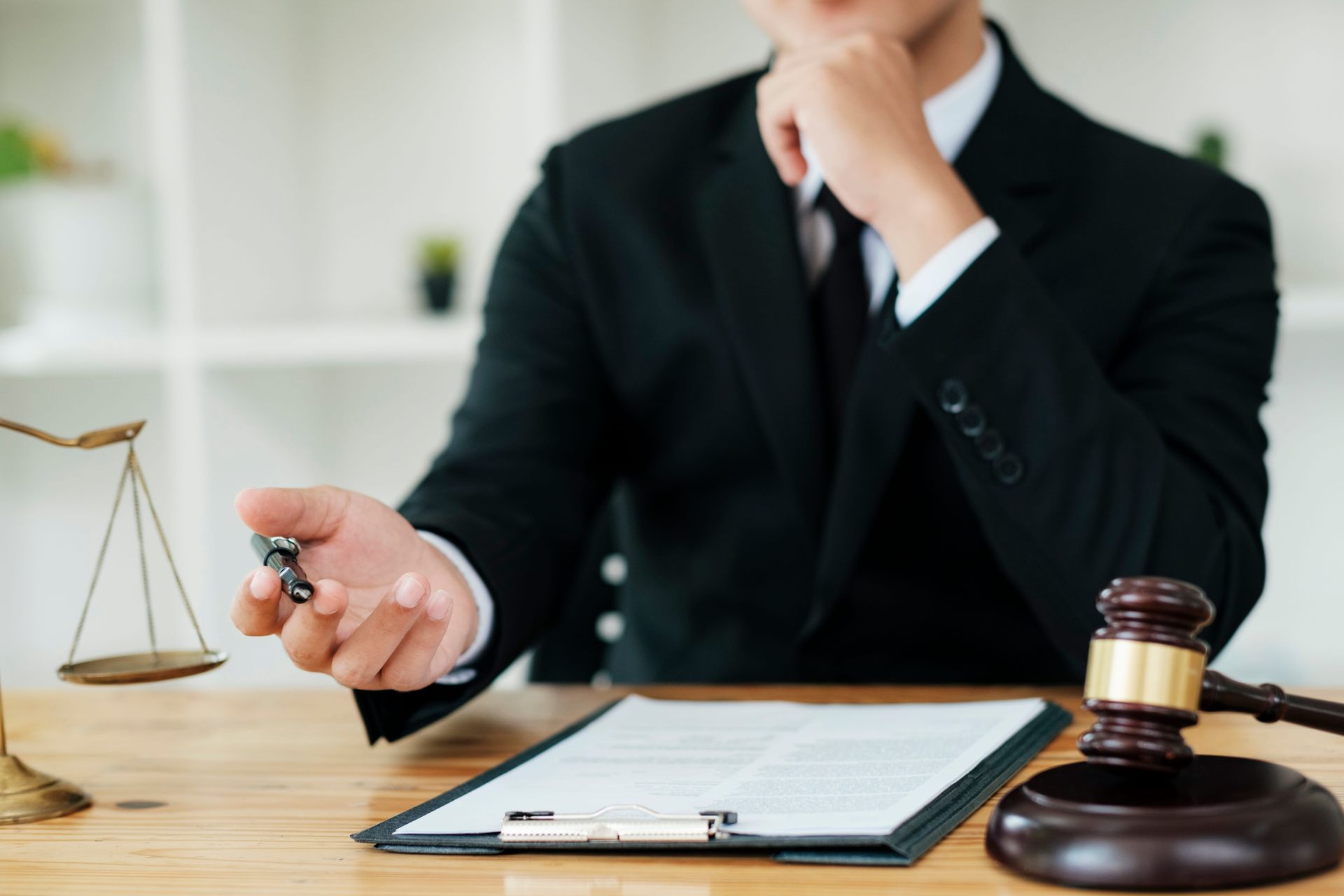 A professional in a suit sits at a desk with a gavel, legal scales, and documents, gesturing toward the foreground.