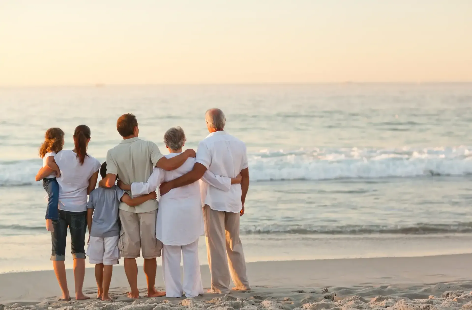 Family of seven standing on beach, arms around each other, looking at the ocean at sunset.