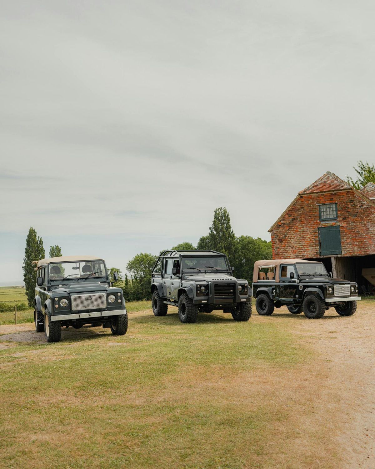 Three Land Rover Defender vehicles parked on a grassy field in front of a brick barn under a cloudy sky.