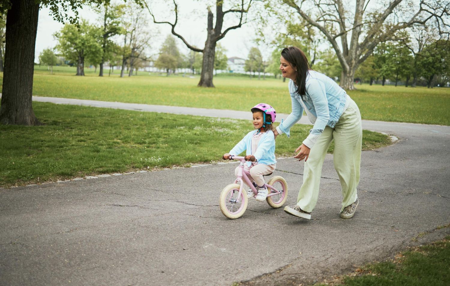 A person helps a young child wearing a pink helmet ride a small bicycle on a paved park path.