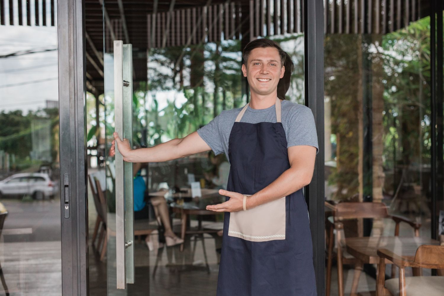 A smiling person in a blue apron holds open a glass door to a restaurant, gesturing for guests to enter.