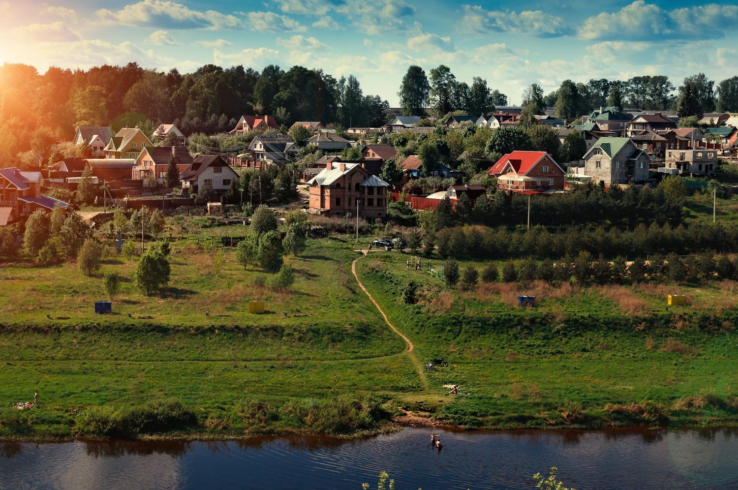 A sunlit landscape shows houses nestled among trees on a hill above a winding river.