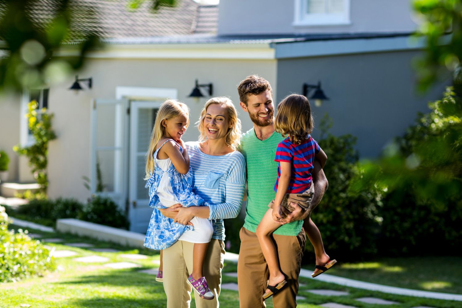 A family of four smiling as they stand together in a sunny garden in front of a house.