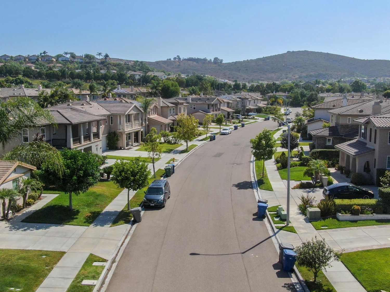 A suburban residential street lined with houses, trees, parked cars, and trash bins under a clear blue sky.