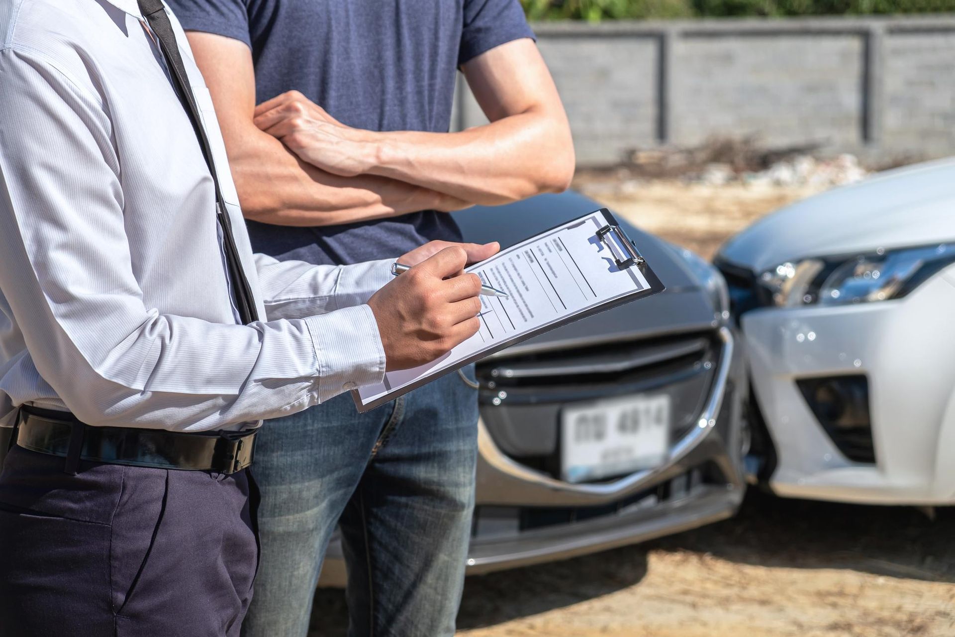 An insurance adjuster writes on a clipboard while standing with a person next to two cars involved in a collision.