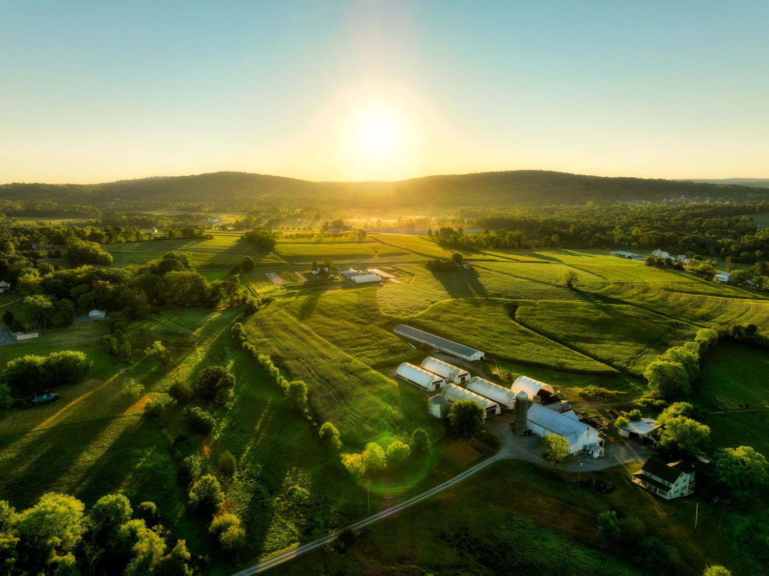 Aerial view of a farm nestled among rolling green hills at sunrise, with light casting long shadows across the landscape.