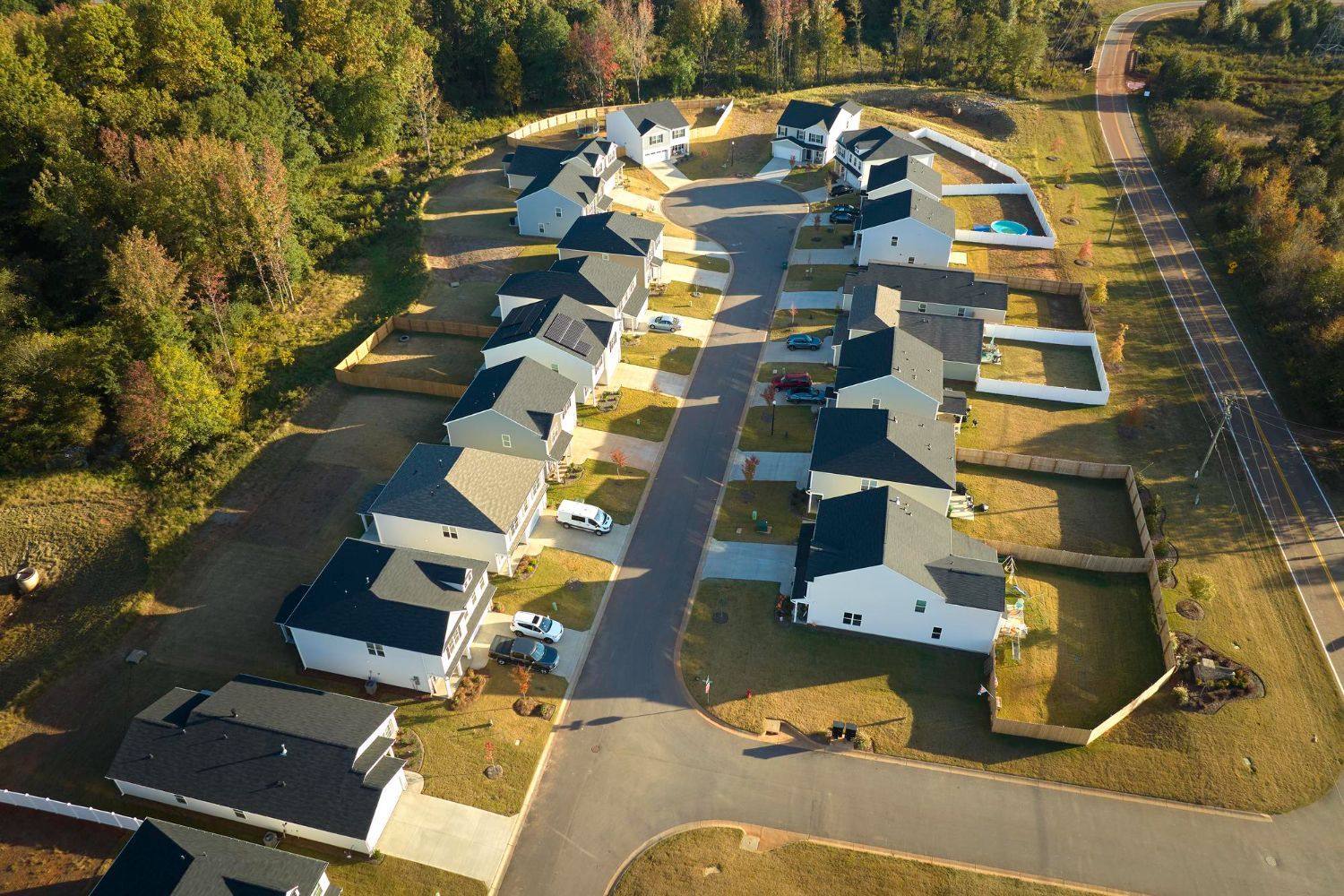 An aerial view of a suburban neighborhood cul-de-sac with white houses, dark roofs, and green lawns surrounded by trees.