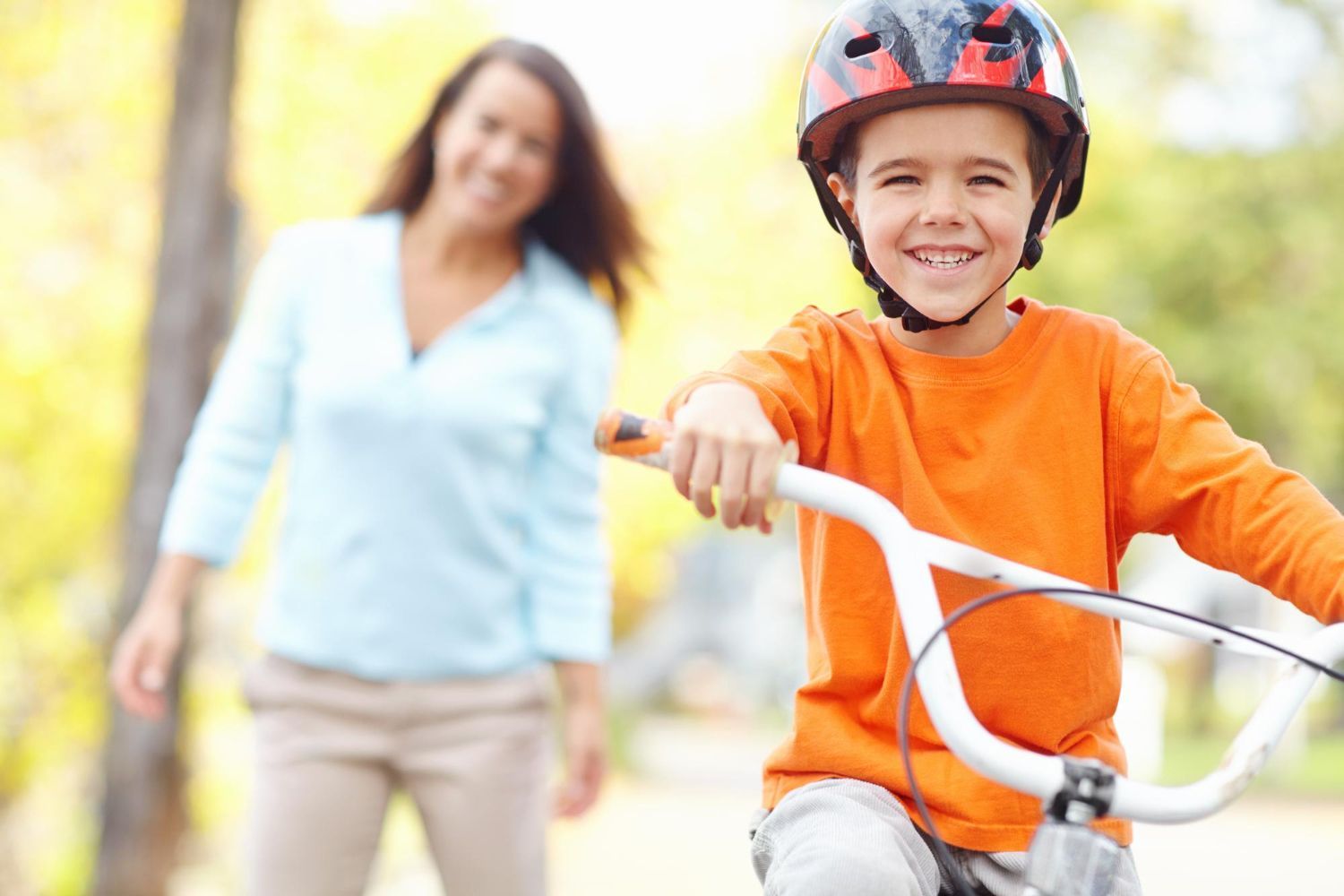 A child wearing a red helmet and orange shirt smiles while riding a bike, with an adult out of focus in the background.