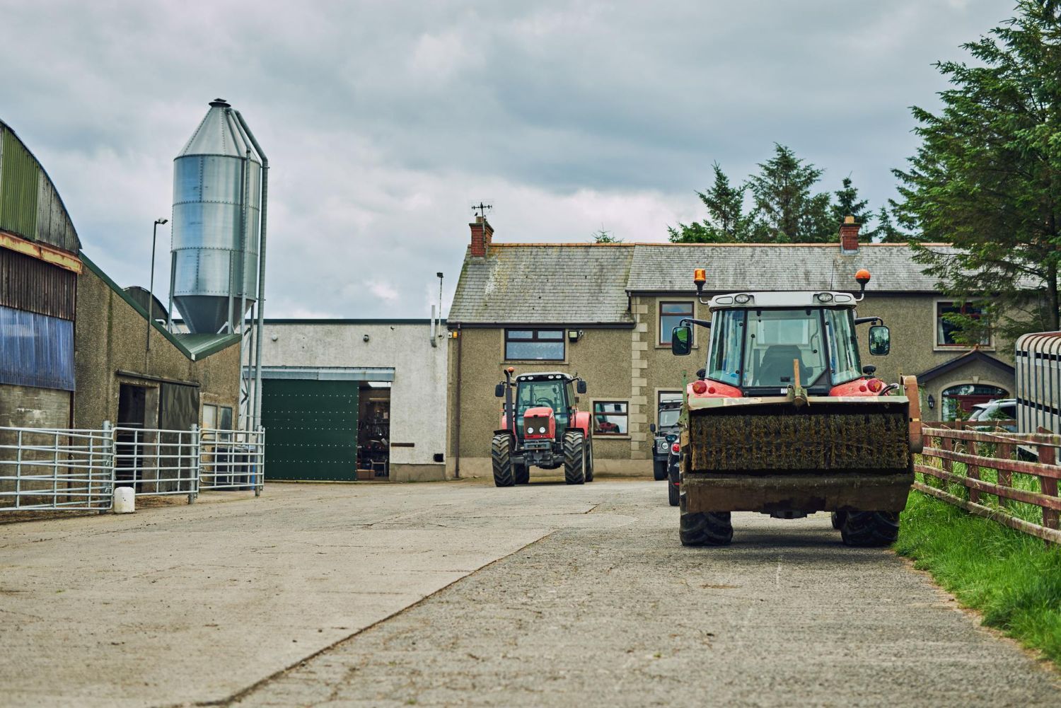 A farmyard with two tractors, a metal grain silo, and farm buildings under a cloudy sky.