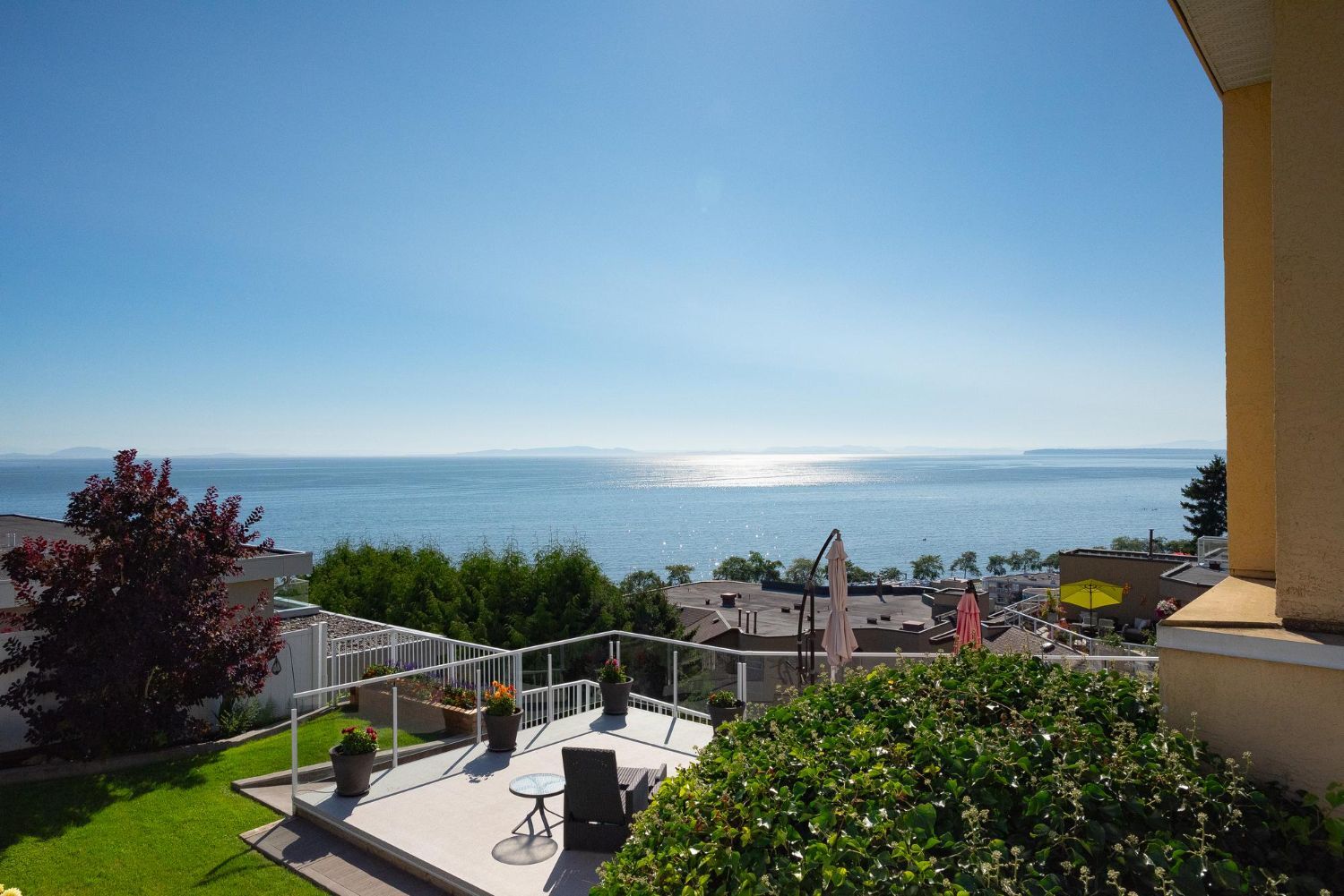 A sunny terrace with patio furniture overlooks a calm sea and distant shoreline under a clear blue sky.