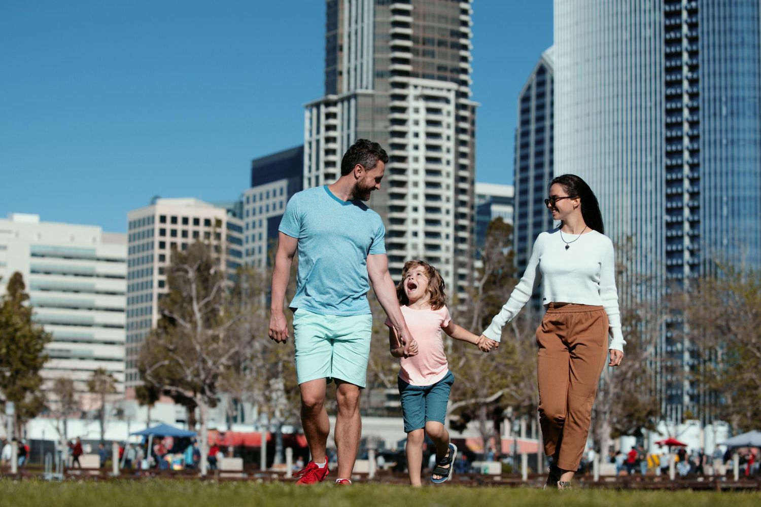 A family walks hand-in-hand through a city park with tall skyscrapers in the background on a sunny day.