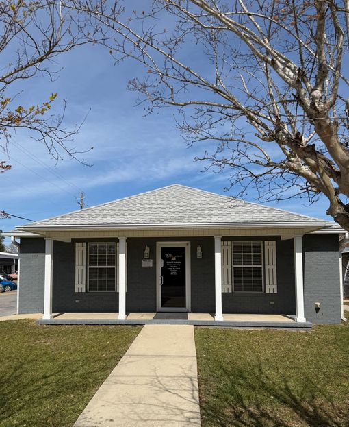 A one-story gray house with a white-pillared front porch, centered walkway, and bare trees under a clear blue sky.