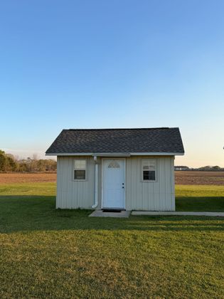 A light-colored shed with a white door, two windows, and a dark shingled roof, situated in a field under a clear blue sky.
