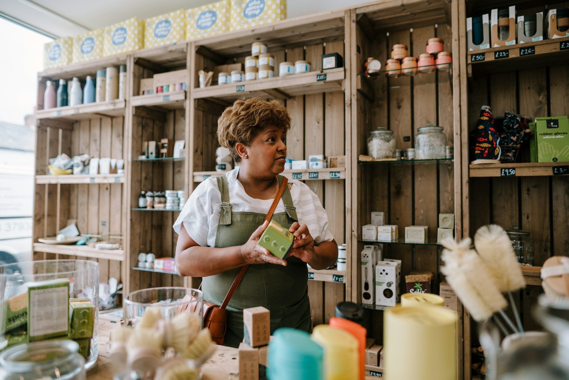 A person wearing olive overalls stands in a shop with wooden shelves, holding a green product in their hands.