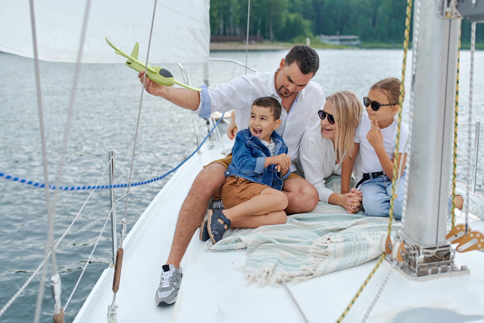 A family of four on a sailboat deck, with a parent holding a toy plane while the children look toward the water.
