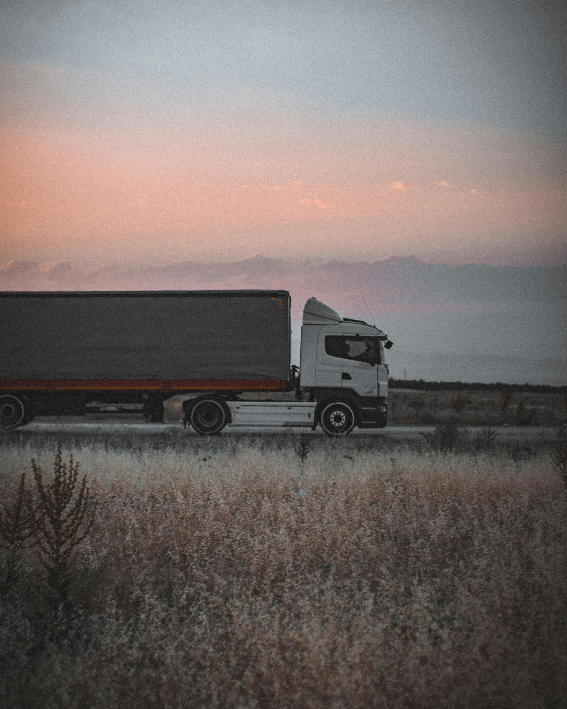 A white semi-truck drives along a road at dusk, seen in profile against a soft pink and gray sky with dry grass in front.