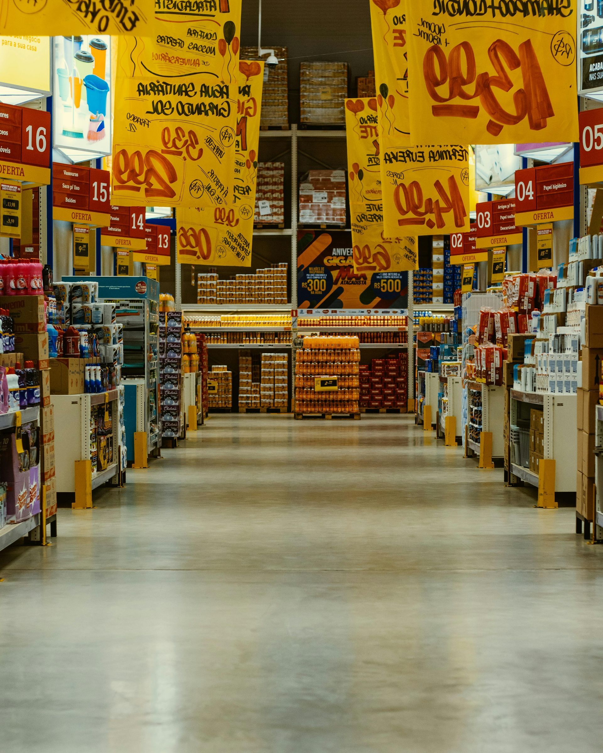 A long, brightly lit aisle in a retail store with yellow promotional banners hanging from the ceiling above shelves.
