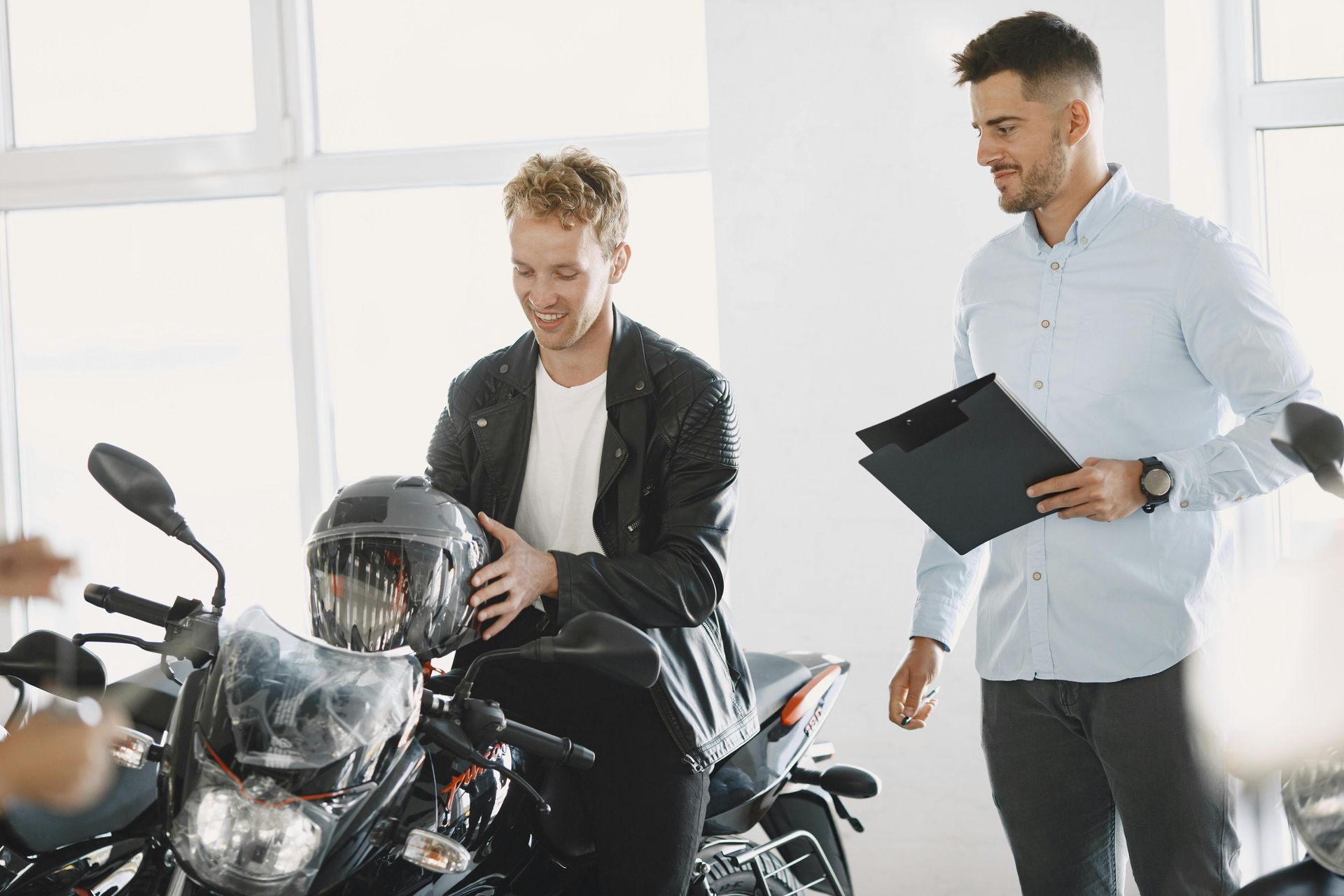 A motorcycle salesperson in a light shirt holding a folder talks to a customer sitting on a bike with a helmet.