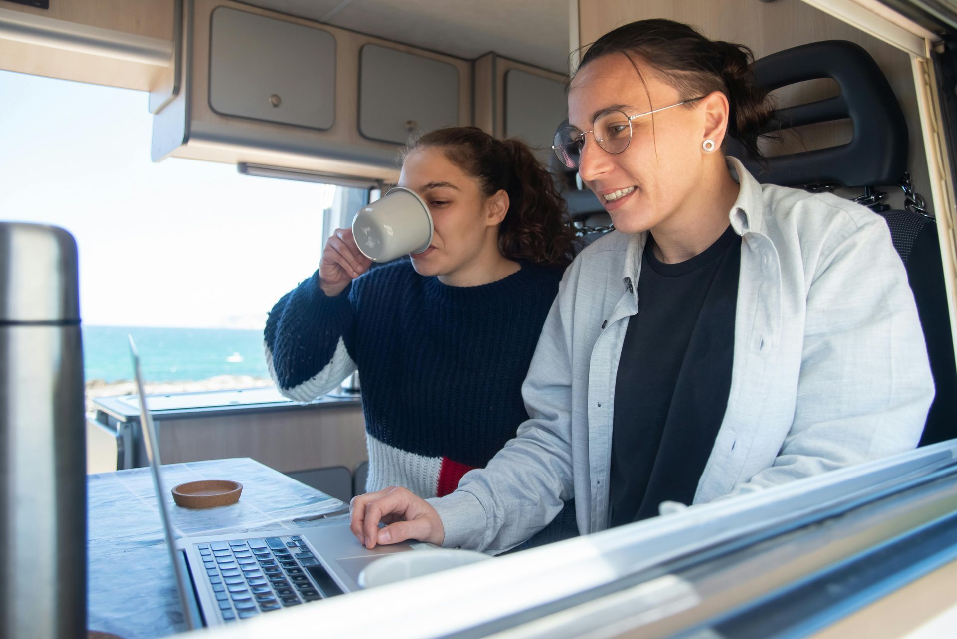 Two people work on a laptop inside a camper van with a view of the ocean, while one drinks from a mug.