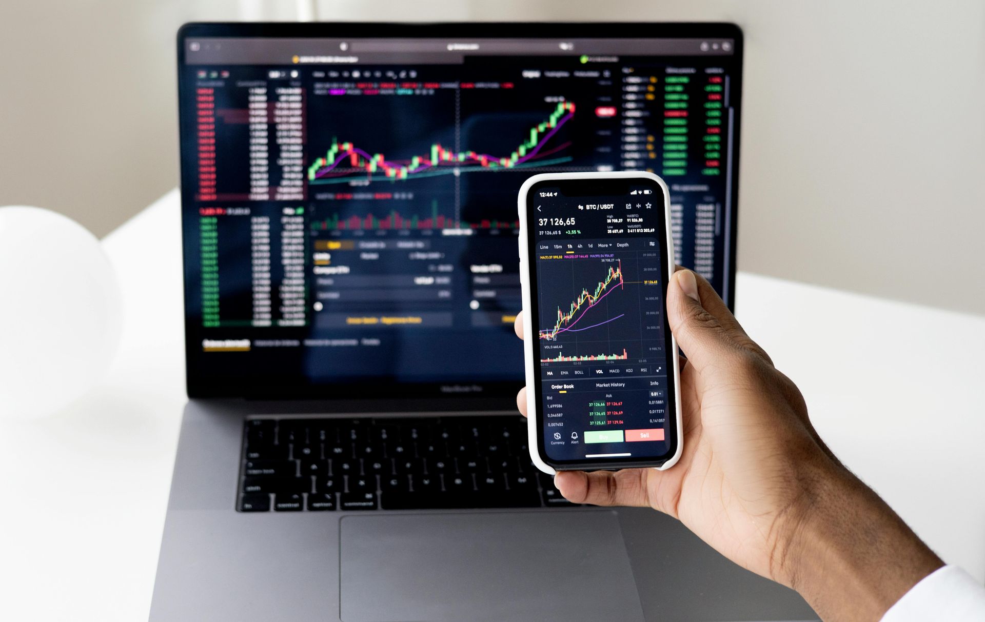 Hand holding a smartphone showing stock charts in front of a laptop with trading graphs on screen