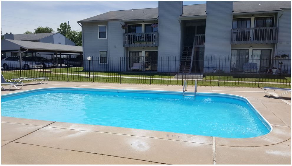 Pool in front of a two-story apartment building with balconies, fenced. Carport and cars visible to the left.