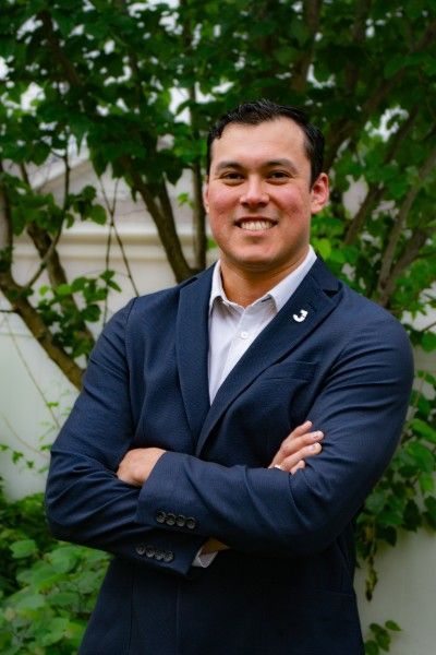 Man with crossed arms smiling, wearing a navy blazer and white shirt, outdoor setting.