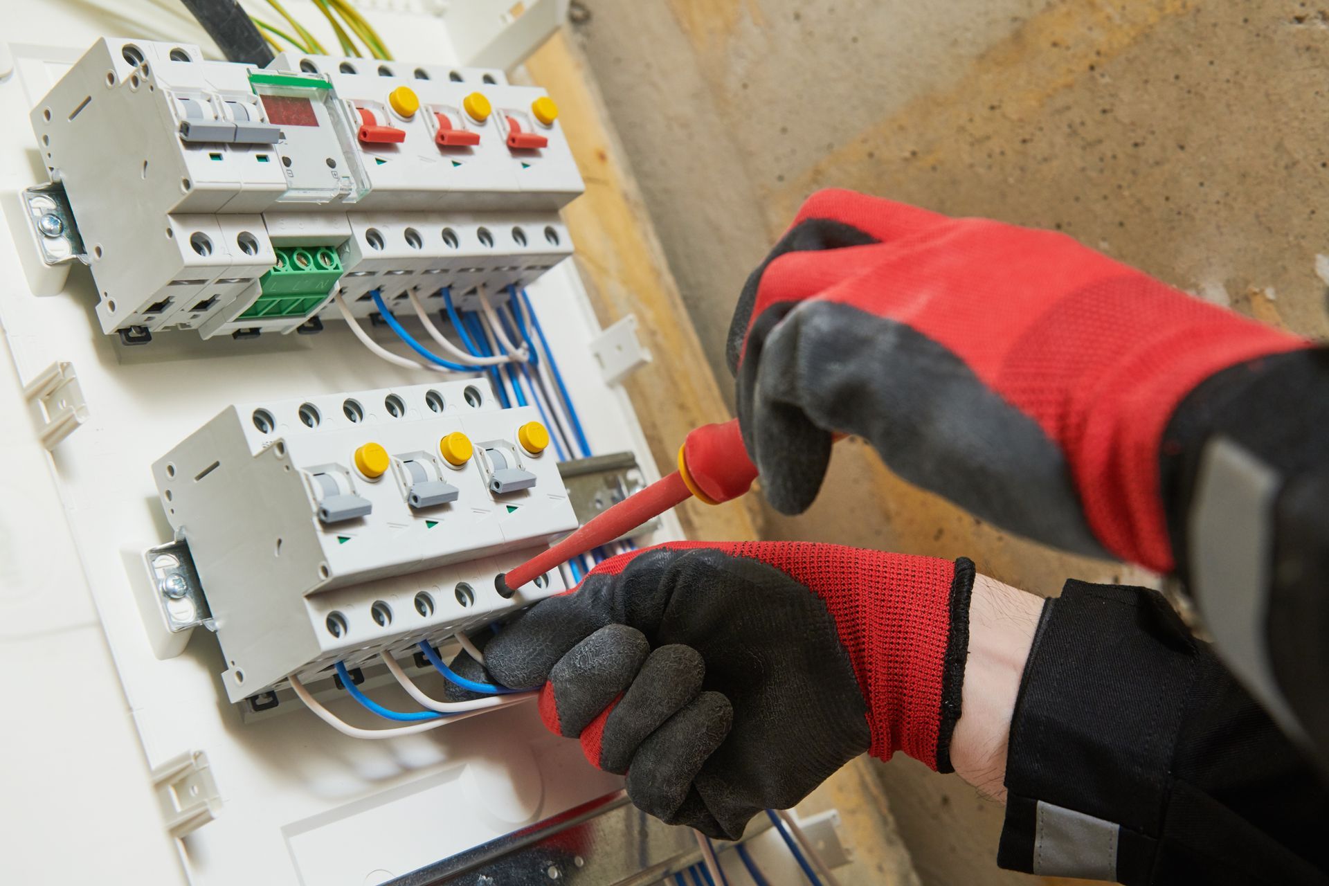 A worker wearing protective gloves uses a screwdriver to adjust components inside an electrical circuit breaker panel.