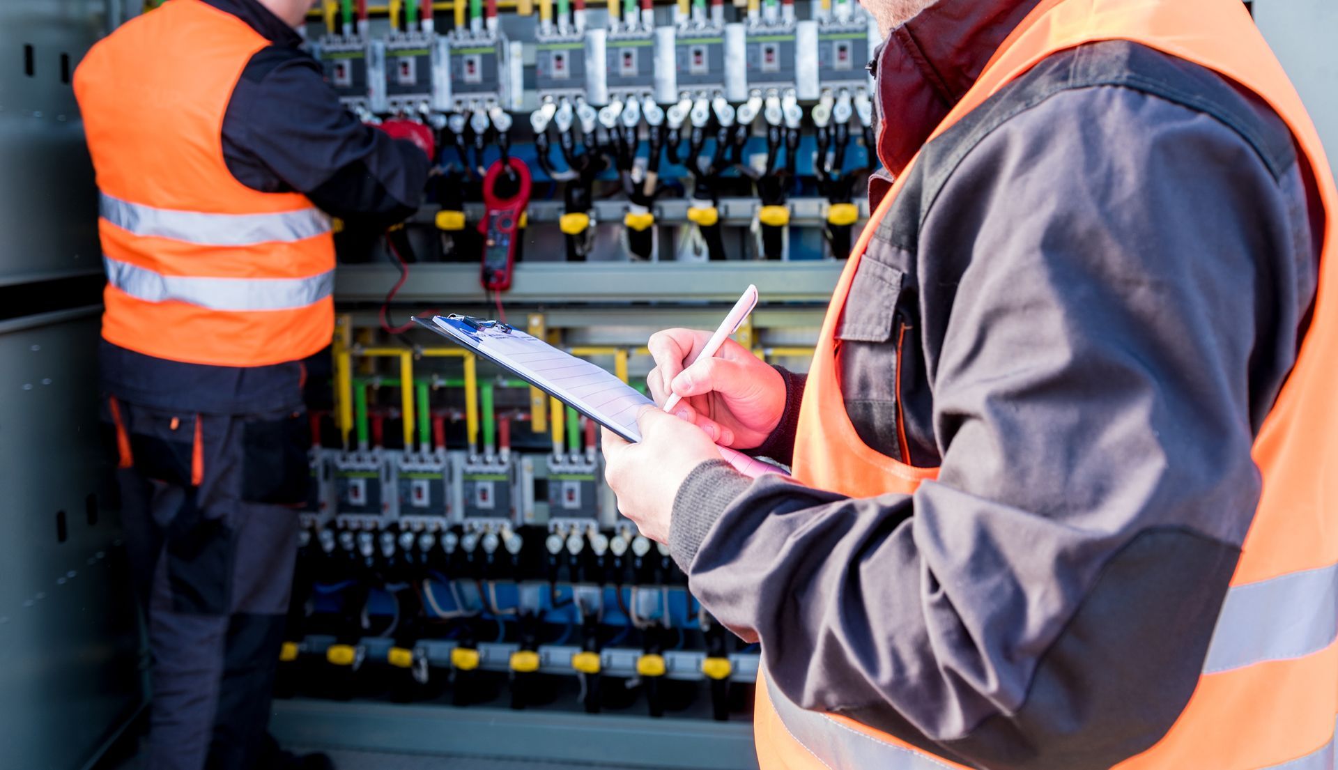 Two technicians in high-visibility orange vests inspect an electrical panel.