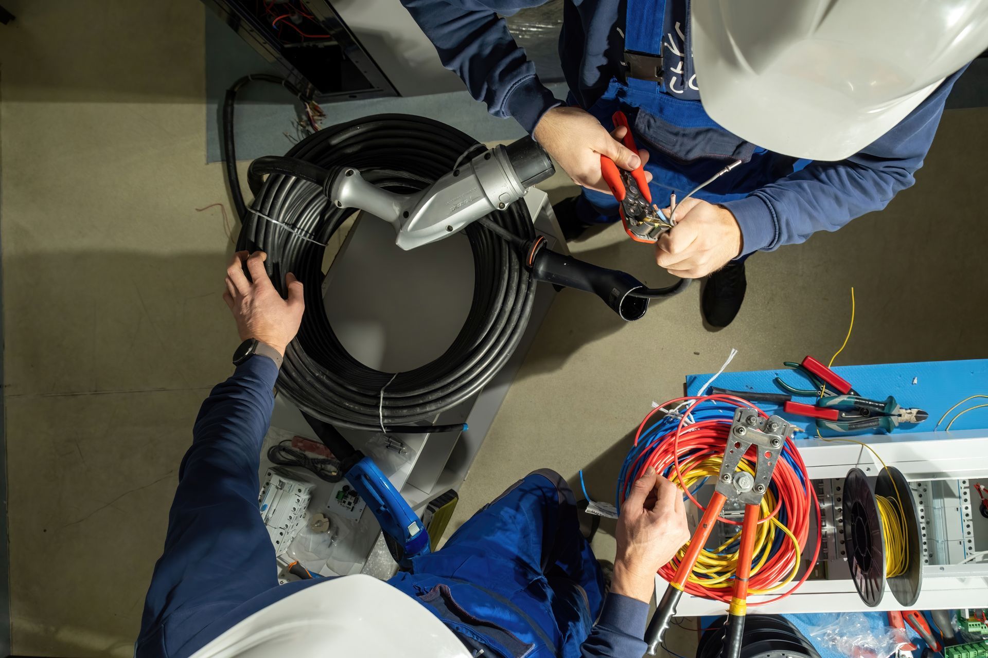 Two workers in blue uniforms and hard hats work with electrical wiring and tools on a floor in an industrial setting.