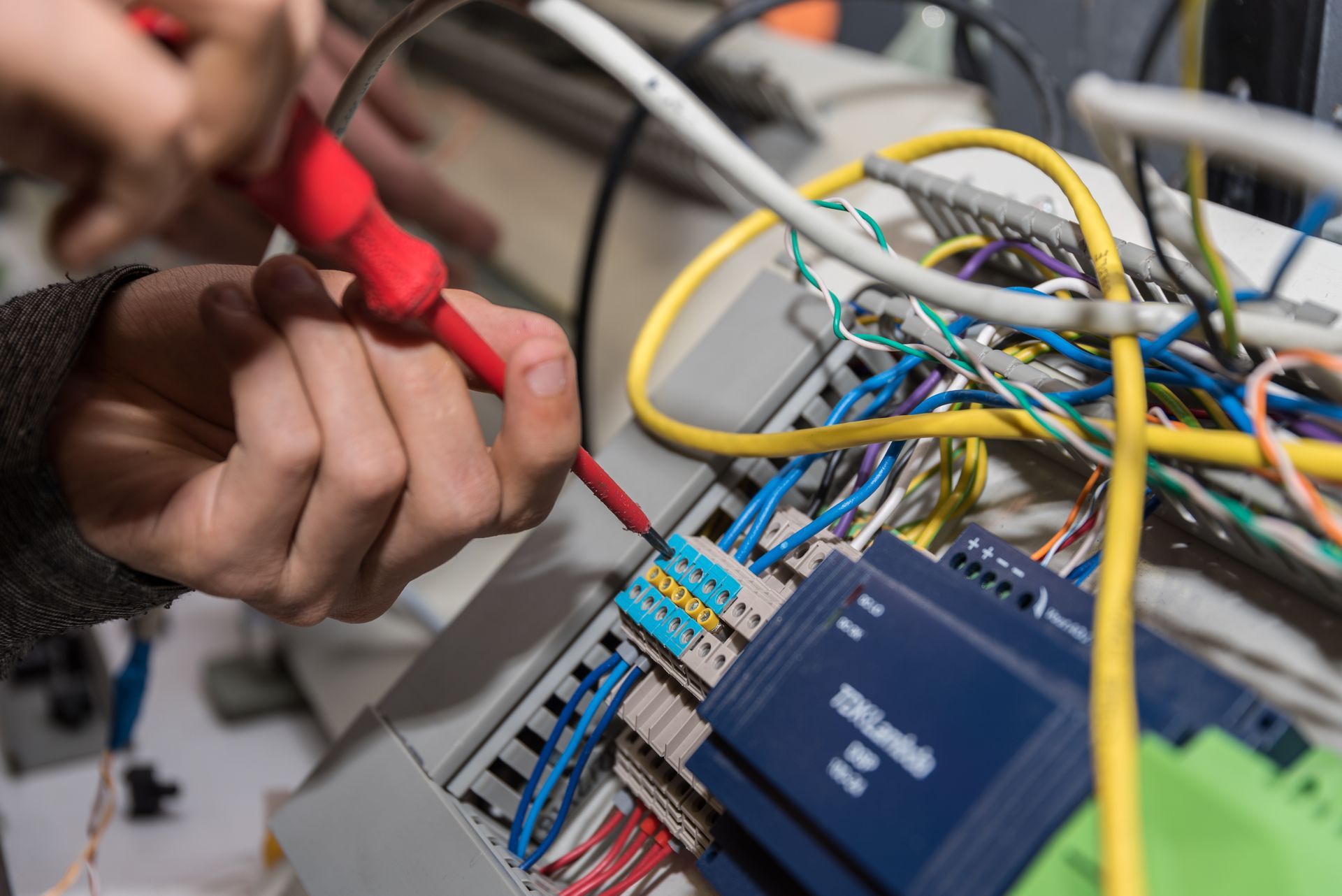A person uses a red screwdriver to secure electrical wires into a blue terminal block in a wiring panel.