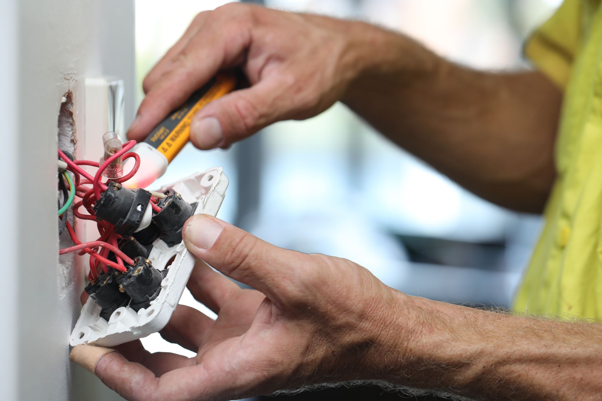 An electrician in a yellow shirt uses a voltage tester to check wires behind a light switch on a wall.