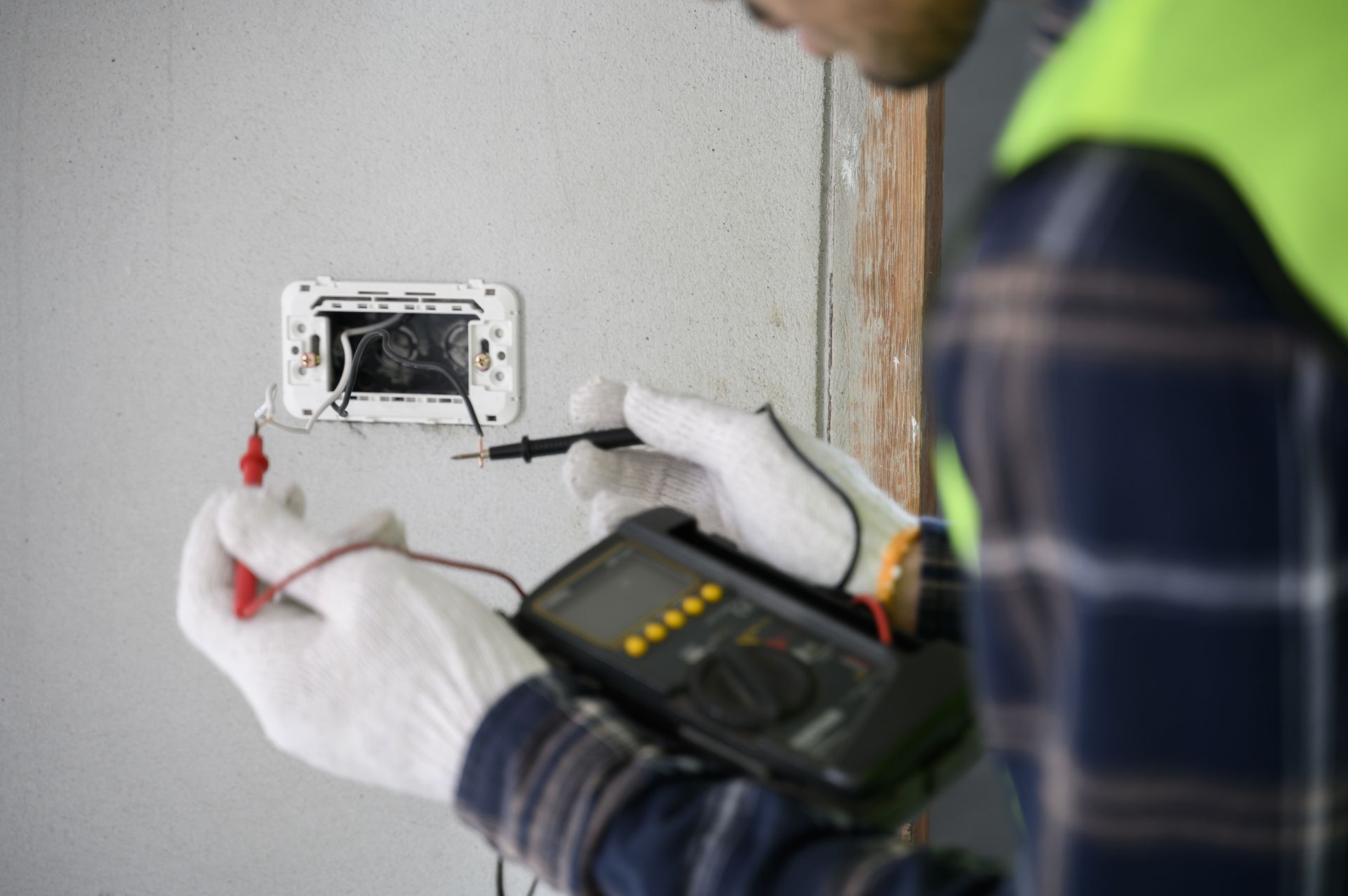 An electrician wearing white gloves uses a multimeter to test electrical wires inside a wall-mounted outlet box.