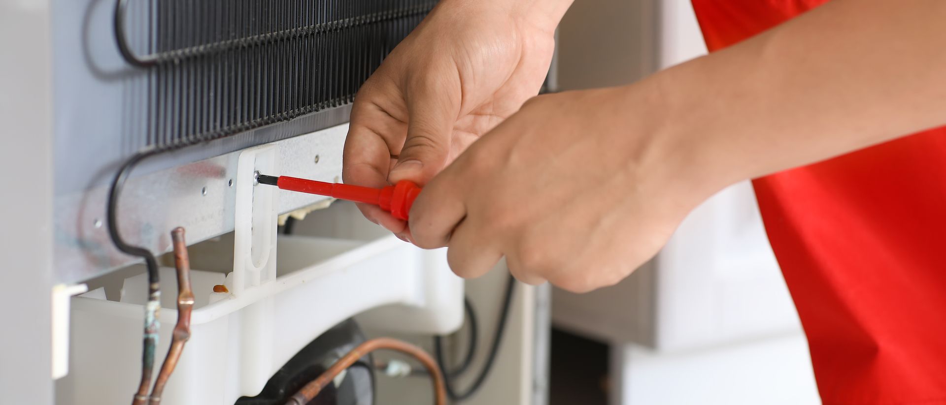 Close-up of a person in a red shirt using a red screwdriver to repair the rear components of a refrigerator.