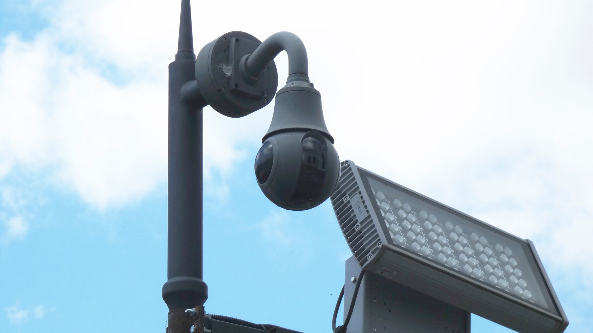 A security camera mounted on a pole next to a rectangular LED floodlight against a cloudy sky.