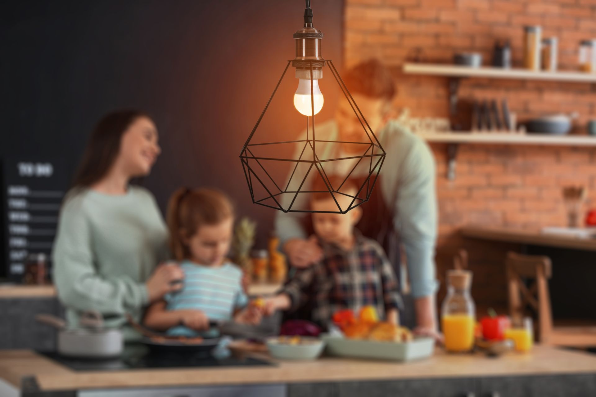A geometric pendant light hangs in the foreground of a blurred kitchen scene with a family preparing a meal together.